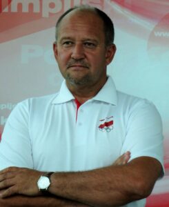 Jacek Puciło szkoleniowiec pływackiej sekcji AZS UWM zdjęcie trenera w białej koszuli na tle tablicy sponsorów sportu