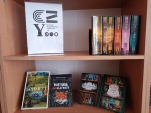 Plakat promujący program Narodowy Program Rozwoju Czytelnictwa 2.0 ns regale obok stoją książki.