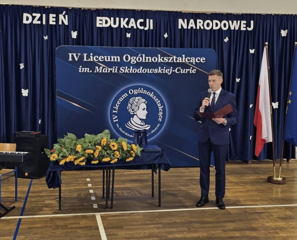Zdjęcie przedstawia dyrektora szkoły, pana Tomasza Niedźwieckiego, w tle zdjęcia ścianka, na której widnieje napis IV Liceum Ogólnokształcące im. Marii Skłodowskiej-Curie z logo szkoły i napis Dzień Edukacji Narodowej.
