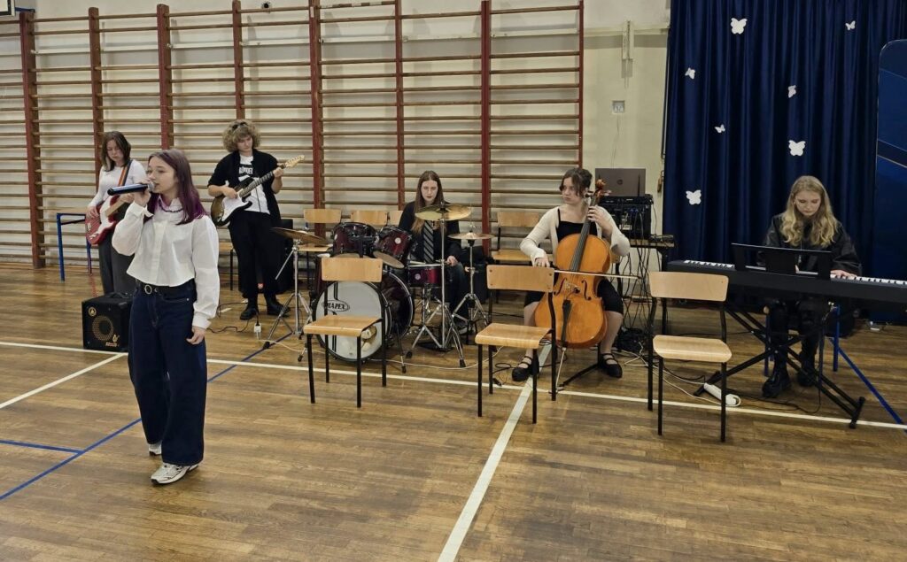 Na zdjęciu zespół muzyczny IV Liceum, grający na instrumentach takich jak gitara, perkusja, pianino, violoncello, na pierwszym planie wokalistka zespołu.